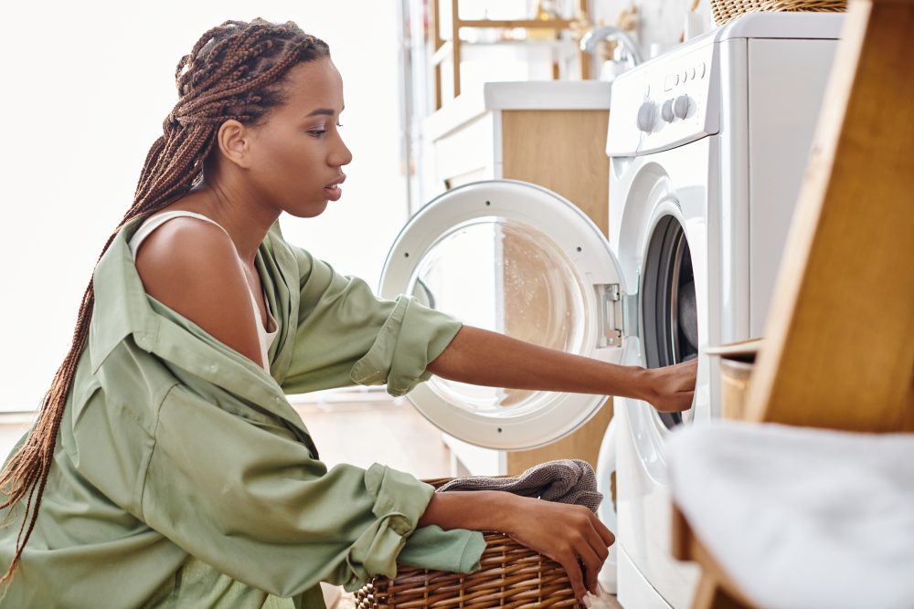 Une femme afro-américaine avec des tresses afro charge une machine à laver dans un sèche-linge tout en faisant la lessive dans une salle de bain.